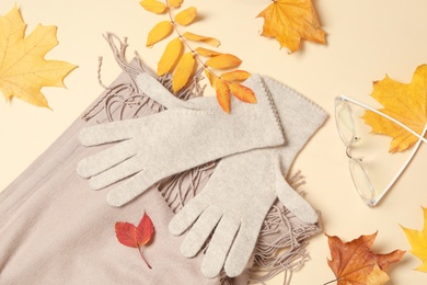 Stylish woolen gloves, scarf, glasses and dry leaves on beige background, flat lay