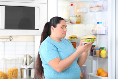 Photo of Sad overweight woman taking sandwich from refrigerator in kitchen. Failed diet