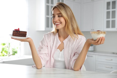 Woman choosing between yogurt with granola and cake at white table in kitchen