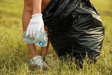 Woman in gloves with trash bag picking crumpled bottle from grass outdoors, closeup