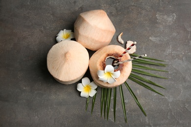 Young peeled coconuts with straw, palm leaf and beautiful flowers on grey table, flat lay