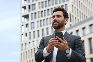 Handsome businessman with smartphone near buildings, space for text