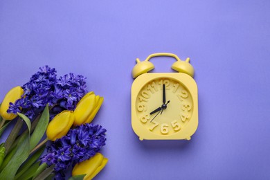 Photo of Yellow alarm clock and beautiful flowers on violet background, flat lay. Spring time