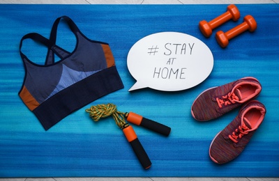 Sport equipment and speech bubble with hashtag STAY AT HOME on blue yoga mat, flat lay. Message to promote self-isolation during COVID‑19 pandemic