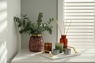 Eucalyptus branches, aromatic reed air freshener and candles on white table near window indoors. Interior element