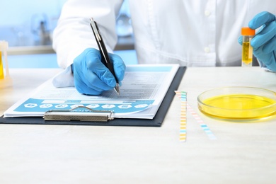 Laboratory assistant with urine sample for analysis writing results of tests at table indoors, closeup