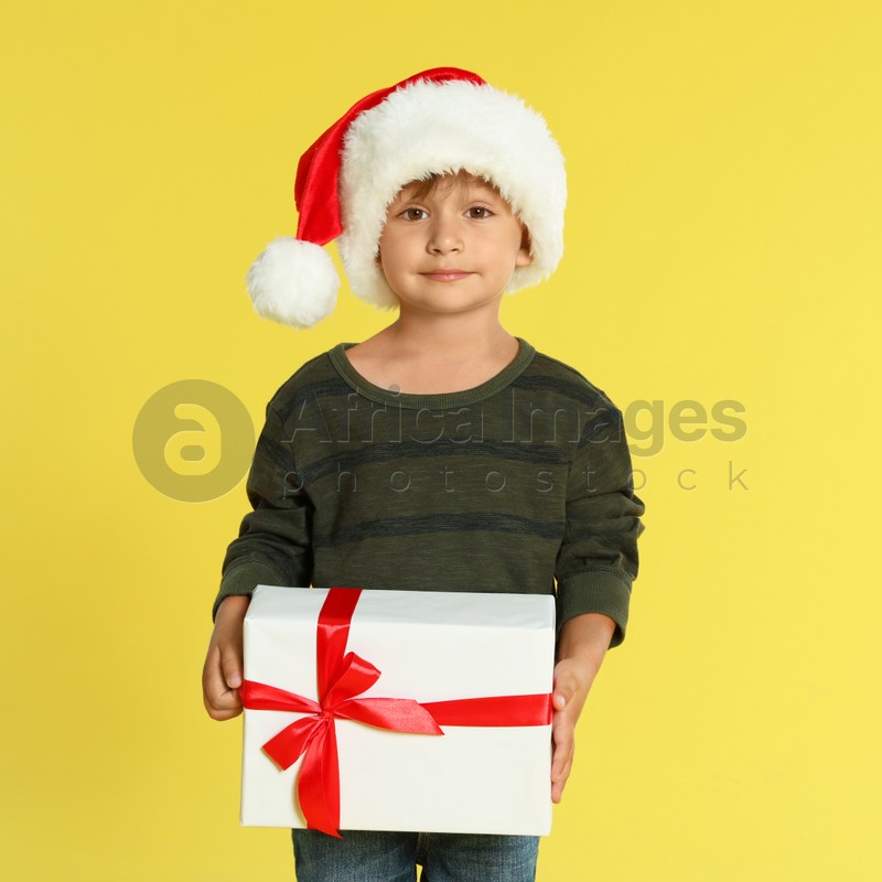 Cute little boy in Santa hat with Christmas gift on yellow background