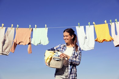 Photo of Smiling woman holding basket with baby clothes near washing line for drying against blue sky outdoors