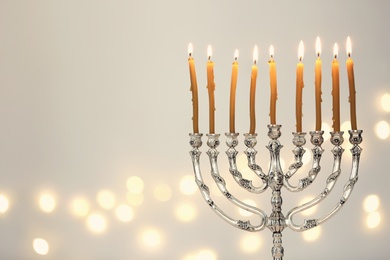 Silver menorah with burning candles against light grey background and blurred festive lights, space for text. Hanukkah celebration