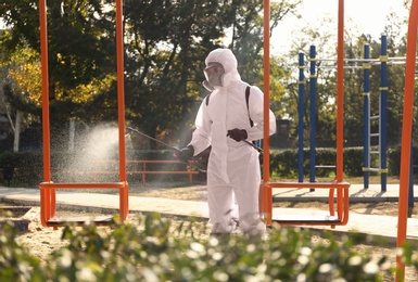 Man in hazmat suit spraying disinfectant onto swing at children's playground. Surface treatment during coronavirus pandemic