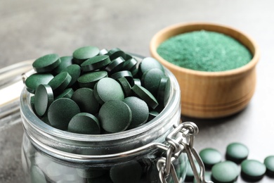 Glass jar with green spirulina pills on grey table, closeup
