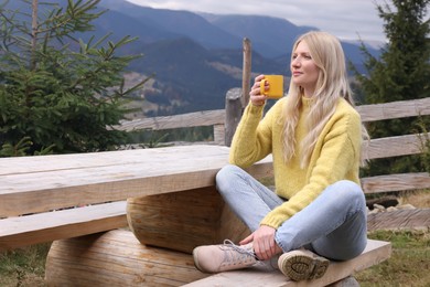 Young woman with mug of hot drink on wooden bench in mountains