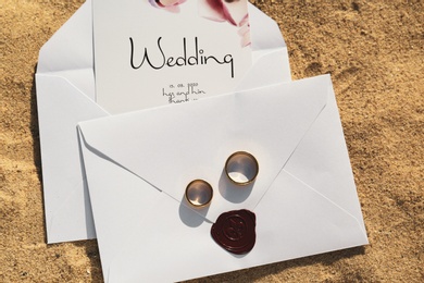 Envelopes with wedding invitations and gold rings on sandy beach, top view