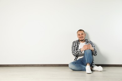Handsome young man sitting on floor near white wall indoors, space for text