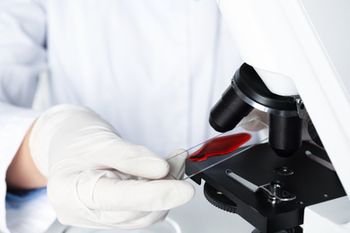 Scientist holding slide with blood sample near microscope in laboratory, closeup. Virus research