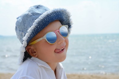 Little girl in sunglasses and hat showing tongue at beach on sunny day. Space for text