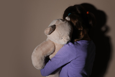 Sad little girl with teddy bear near beige wall, back view. Domestic violence concept