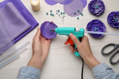 Woman with hot glue gun making textile flower at white wooden table, top view