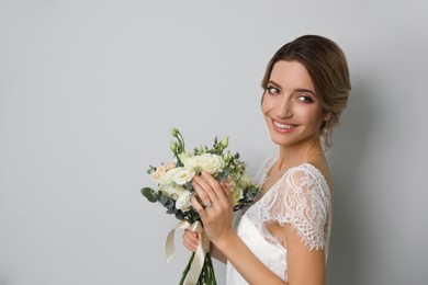 Young bride with elegant hairstyle holding wedding bouquet on light grey background