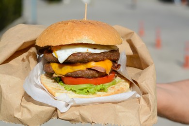 Photo of Woman holding delicious burger in paper wrap outdoors, closeup