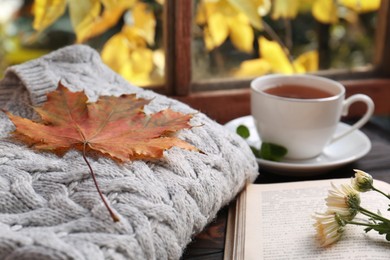 Photo of Cup of aromatic tea, soft sweater and book on wooden windowsill indoors, closeup. Autumn atmosphere