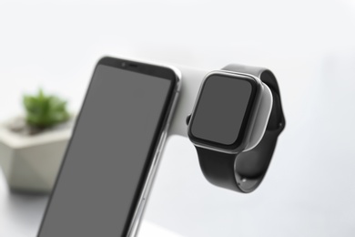 Stylish smart watch and phone charging with wireless pad against blurred background, closeup