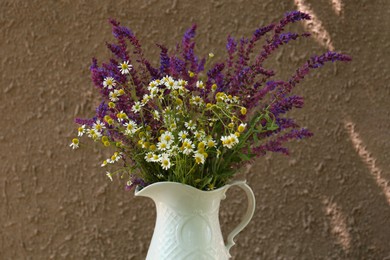 Beautiful bouquet with field flowers in jug near brown wall