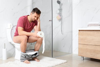 Man with paper roll suffering from stomach ache on toilet bowl in bathroom
