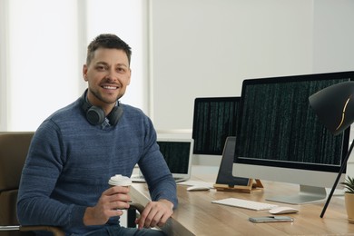 Happy programmer with headphones and coffee working at desk in office