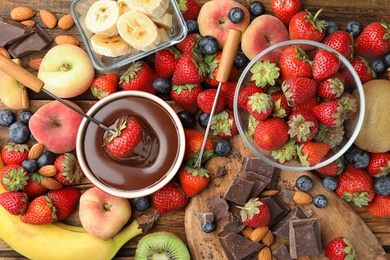Fondue fork with strawberry in bowl of melted chocolate surrounded by other fruits on wooden table, flat lay