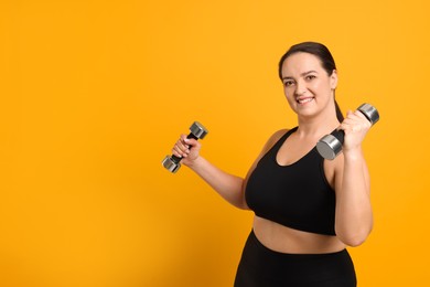 Happy overweight woman doing exercise with dumbbells on orange background, space for text