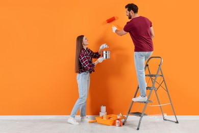 Photo of Man painting orange wall and happy woman holding can of paint indoors. Interior design