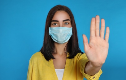 Young woman in protective mask showing stop gesture on light blue background. Prevent spreading of coronavirus