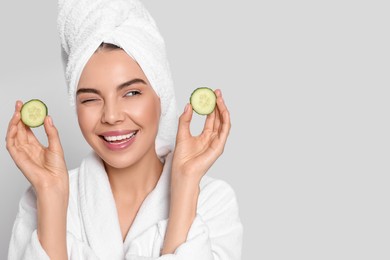Photo of Woman in bathrobe with towel holding pieces of cucumber on light grey background, space for text. Spa treatment