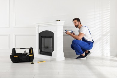 Professional technician sealing electric fireplace with caulk near white wall in room