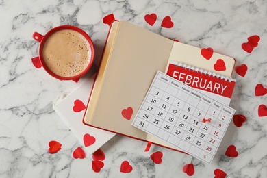 Calendar with marked Valentine's Day, notebooks and cup of coffee on white marble table, flat lay