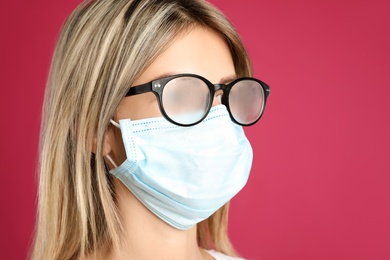 Woman with foggy glasses caused by wearing disposable mask on pink background. Protective measure during coronavirus pandemic