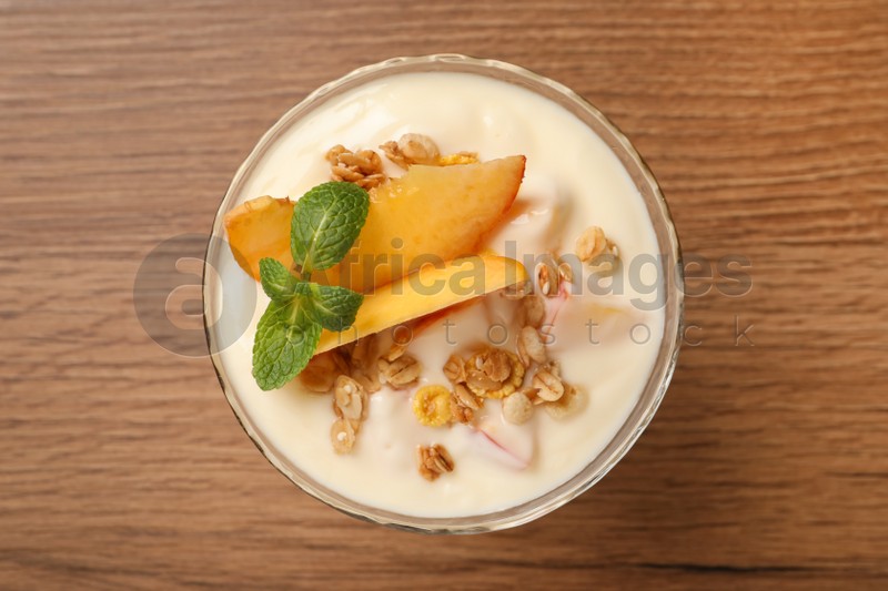 Tasty peach yogurt with granola, mint and pieces of fruit in dessert bowl on wooden table, top view