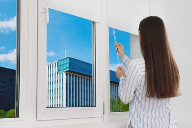 Photo of Woman opening white roller blinds on window indoors