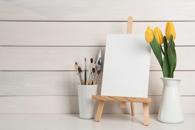 Easel with blank canvas, brushes and tulips on white wooden table. Space for text