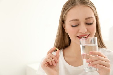 Photo of Young woman with glass of water taking vitamin capsule on light background. Space for text