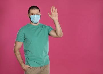 Man in protective mask showing hello gesture on pink background, space for text. Keeping social distance during coronavirus pandemic