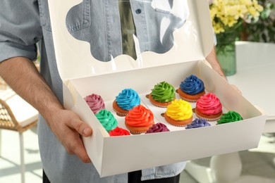 Man holding box with delicious colorful cupcakes indoors, closeup