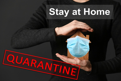 Stay at home during coronavirus quarantine. Woman holding wooden house model with medical mask on dark background