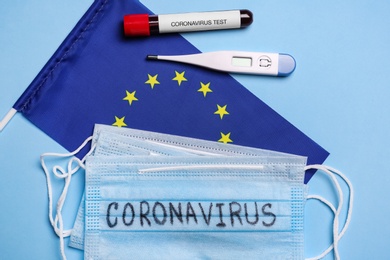 European Union flag, protective masks, thermometer and test tube with blood sample on light blue background, flat lay. Coronavirus outbreak