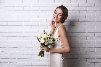 Young bride with elegant hairstyle holding wedding bouquet near white brick wall