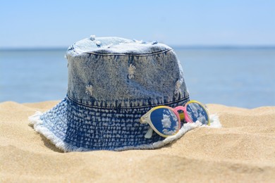 Photo of Jeans hat and sunglasses on sand near sea, closeup. Beach accessories