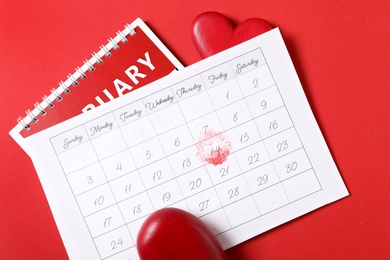 Calendar with marked Valentine's Day and wooden hearts on red background, flat lay