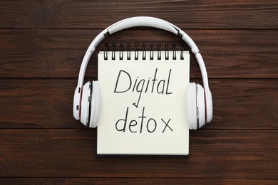 Notebook with phrase DIGITAL DETOX and headphones on wooden table, flat lay