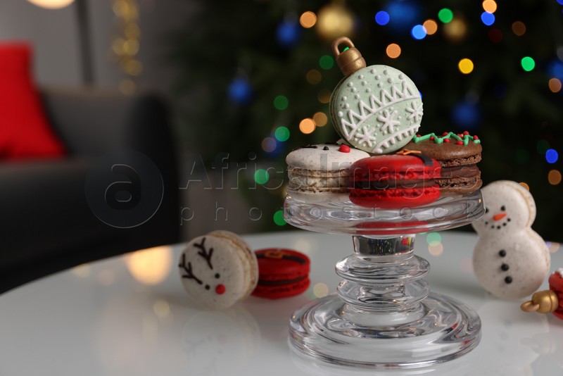 Beautifully decorated Christmas macarons on white table against blurred festive lights, space for text